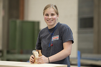 Zimmerin bei ZÜBLIN Timber Junge Frau an einer Werkbank, lächelt in die Kamera.