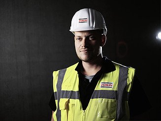 Newcomer Portraitfoto eines Poliers mit Warnweste und Bauhelm.