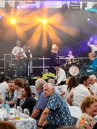 Bühne mit Liveband, im Vordergrund sitzen Personen auf Bierbänken.