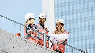 Foto von zwei Personen auf Baustelle, wobei die Frau nach oben zeigt 