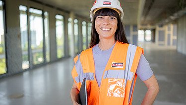 Foto von einer lachenden ZÜBLIN-Architektin in Arbeitssicherheitskleidung auf einer Baustelle mit Tablet im Arm.