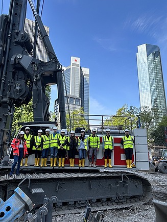 Kinder in Arbeitsschutzkleidung stehen auf einer Baustelle auf einem Riesenbohrgerät, im Hintergrund die Frankfurter Skyline.