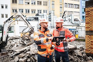 Zwei Mitarbeitende in Arbeitsschutzkleidung auf einer Baustelle im Gespräch, einer mit Tablet