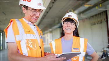 Foto von zwei ZÜBLIN-Mitarbeitenden, die lachend zusammen auf der Baustelle stehen und etwas besprechen.