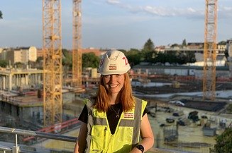 Junge Frau in Arbeitskleidung auf einer Baustelle, lächelt in die Kamera.