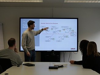 Teammeeting mit mehreren Personen, ein Kollege steht vorne und präsentiert