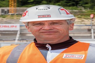 Portrait von einem Mann in Arbeitsschutzkleidung, im Hintergrund ist eine Baustelle zu sehen