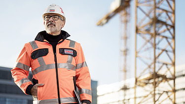 A photo of a ZÜBLIN quantity surveyor on a building site, wearing PPE, looking towards the camera.