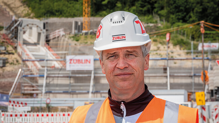 Foto von einem Mann in Arbeitsschutzkleidung, im Hintergrund eine Baustelle