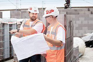 Zwei Mitarbeiter in Arbeitsschutzkleidung mit einem Bauplan auf der Baustelle.