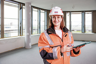 A young woman in high-visibility workwear holding a tablet on a building site.