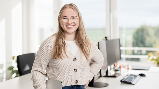Junge Frau lächelt an ihrem Büro-Arbeitsplatz in die Kamera.