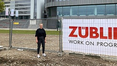 Junge Frau steht vor einem Bauzaun, im Hintergrund ist die Uni Bielefeld zu sehen