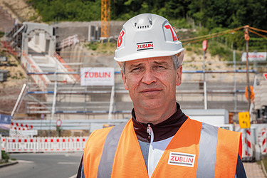 Foto von einem Mann in Arbeitsschutzkleidung, im Hintergrund eine Baustelle