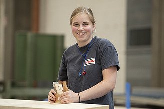 Junge Frau an einer Werkbank, lächelt in die Kamera.