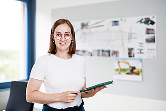 A young woman is standing in an office holding a tablet and smiling at the camera.