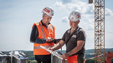 Zwei Männer in Arbeitsschutzkleidung im Gespräch, im Hintergrund ein Kran