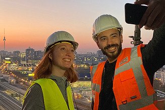 Selfie einer jungen Frau und eines jungen Mannes in Arbeitskleidung auf einer Baustelle, im Hintergrund ist der Sonnenuntergang über Berlin zu sehen.
