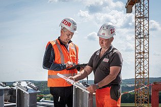Zwei Männer in Arbeitsschutzkleidung im Gespräch, im Hintergrund ein Kran