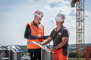 Two men in high-visibility workwear talking, with a crane in the background