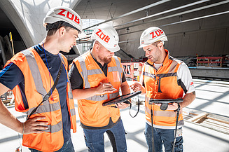A photograph of three foremen standing in front of a building plan and discussing something.