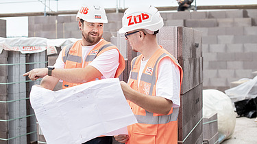 Zwei Mitarbeiter in Arbeitsschutzkleidung mit einem Bauplan auf der Baustelle.