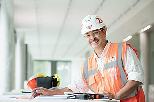 A photograph of a ZÜBLIN employee standing on a building site, poring over a construction plan and smiling at the camera.