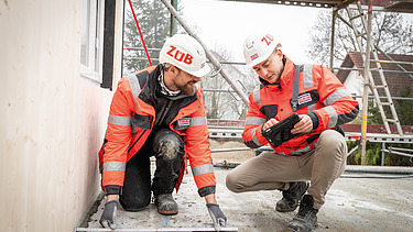 Two staff members in high-visibility clothing talking on a building site; one is holding a tablet, the other a folding rule