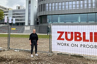 Junge Frau steht vor einem Bauzaun, im Hintergrund ist die Uni Bielefeld zu sehen