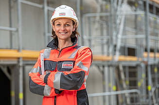 Frau steht mit persönlicher Schutzausrüstung auf einer Baustelle und lächelt in die Kamera.