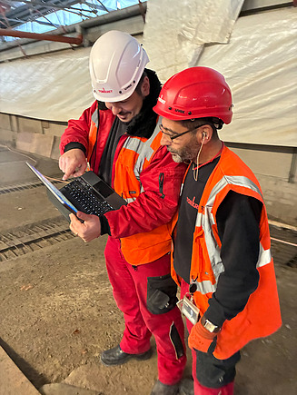 Zwei Männer in Arbeitsschutzkleidung auf einer Baustelle, einer hält ein Tablet in der Hand