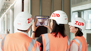 Photo of three ZÜBLIN staff members on a building site, viewing a digital building plan on a tablet.