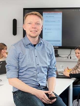 Junger Mann in einem Meetingraum lächelt in die Kamera, im Hintergrund vier weitere Personen an einem Besprechungstisch