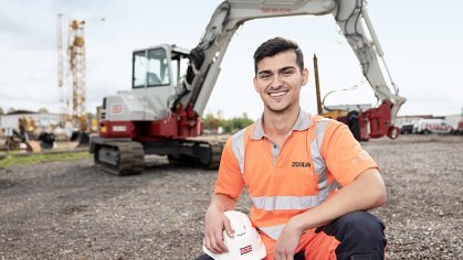 A young man in work clothes, with an excavator in the background.