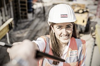 Junge Frau in Arbeitskleidung auf einer Baustelle, lächelt in die Kamera.