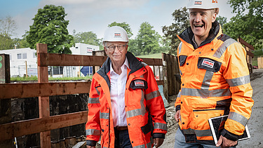 Zwei Mitarbeiter in Arbeitsschutzkleidung auf der Baustelle, lachen in die Kamera