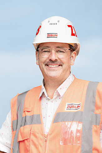 A worker on a building site, wearing a hard hat and a high-visibility vest, smiles for the camera. 