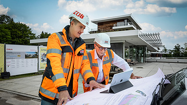 Zwei Mitarbeitende in Arbeitsschutzkleidung über ein Tablet und einen Bauplan gebeugt