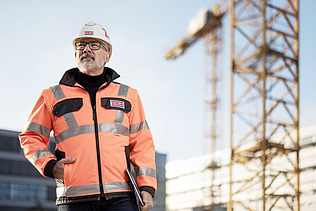 A photo of a ZÜBLIN quantity surveyor on a building site, wearing PPE, looking towards the camera.
