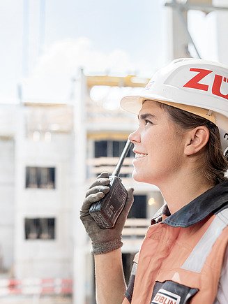 Junge Frau mit Arbeitskleidung auf einer Baustelle, hält Funkgerät in der Hand