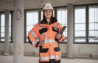 Foto einer ZÜBLIN-Mitarbeiterin, die in Arbeitsschutzkleidung und einem Tablet auf einer Baustelle steht und in die Kamera schaut.
