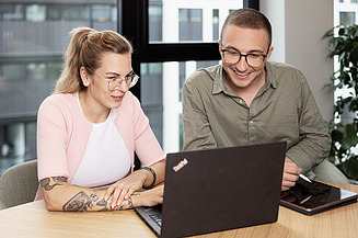 A photo of a smiling woman with her arms crossed in the foreground, whilst two people are talking in the background 