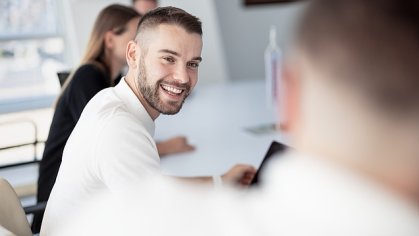 A young man is sitting at a desk in an office and smiling at the camera.