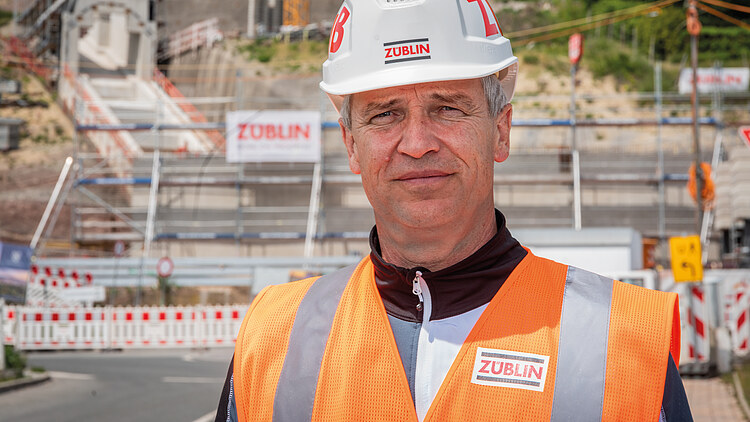 Foto von einem Mann in Arbeitsschutzkleidung, im Hintergrund eine Baustelle