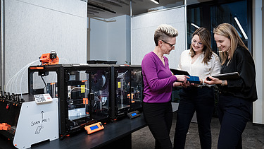 Three ZÜBLIN employees standing in front of a 3D printer and discussing something.