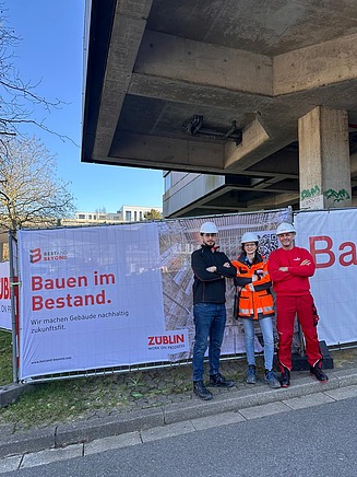 Drei Mitarbeitende in Arbeitsschutzkleidung mit verschränkten Armen vor einem, Bauzaun mit 