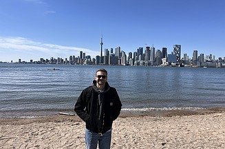 A young man is standing on the riverbank, with the Toronto skyline in the background