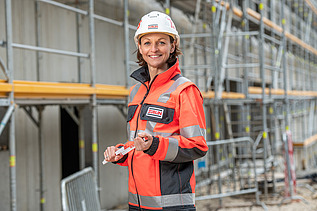 Frau steht mit persönlicher Schutzausrüstung auf einer Baustelle und lächelt in die Kamera.