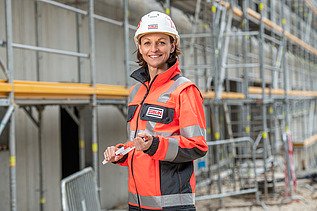 Frau steht mit persönlicher Schutzausrüstung auf einer Baustelle und lächelt in die Kamera.