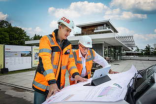 Zwei Mitarbeitende in Arbeitsschutzkleidung über ein Tablet und einen Bauplan gebeugt